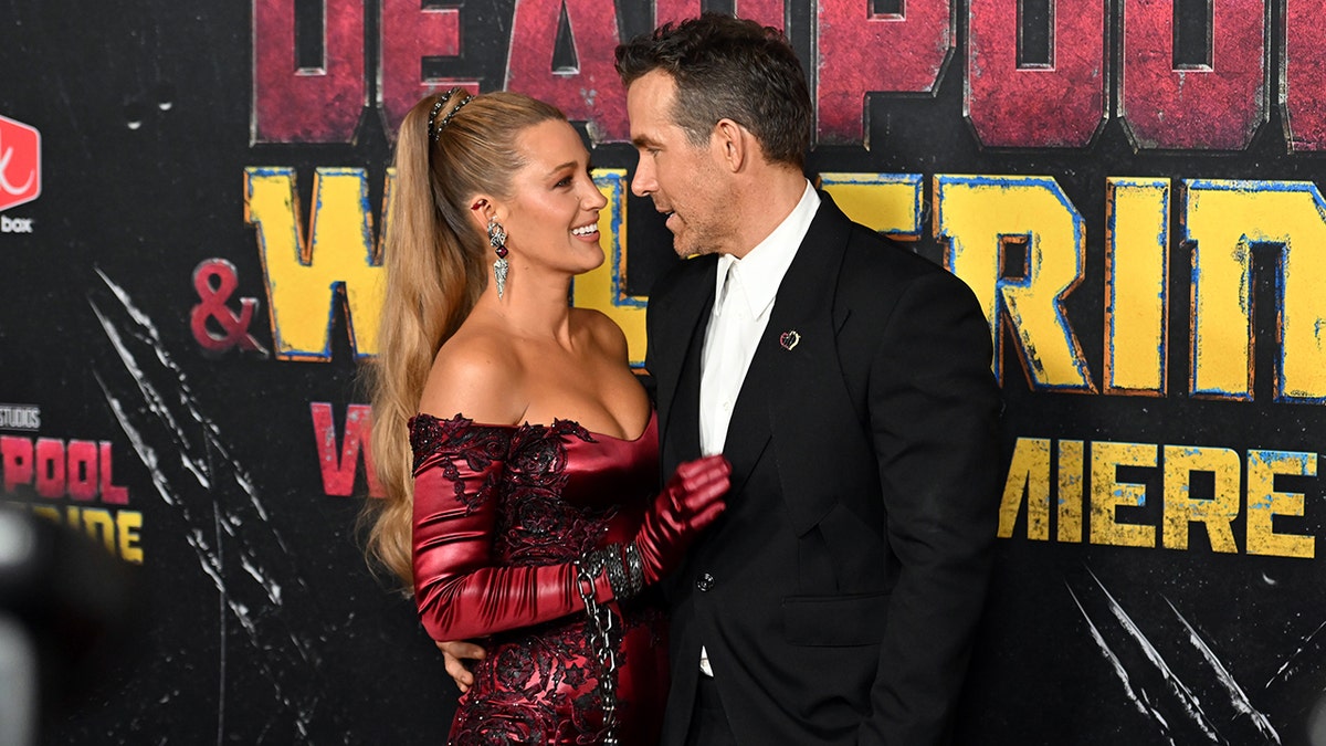 Blake Lively in a red printed jumpsuit holding onto her husband Ryan Reynolds in a black suit and white shirt