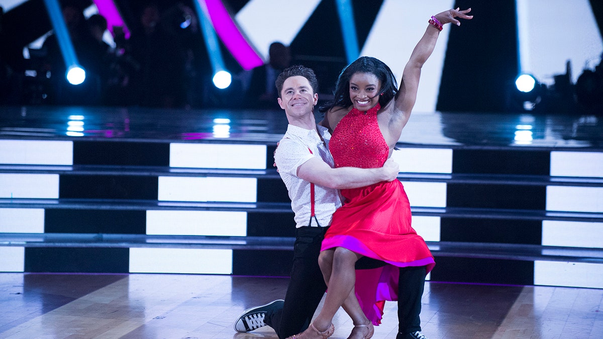 Sasha Farber in a white shirt kneels as Simone Biles in a red dress raises her arm during 'Dancing with the Stars'