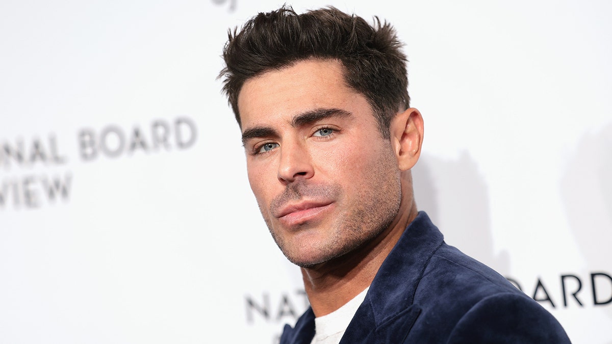 Zac Efron glances toward the camera in a stylish navy velvet suit at a red carpet event.