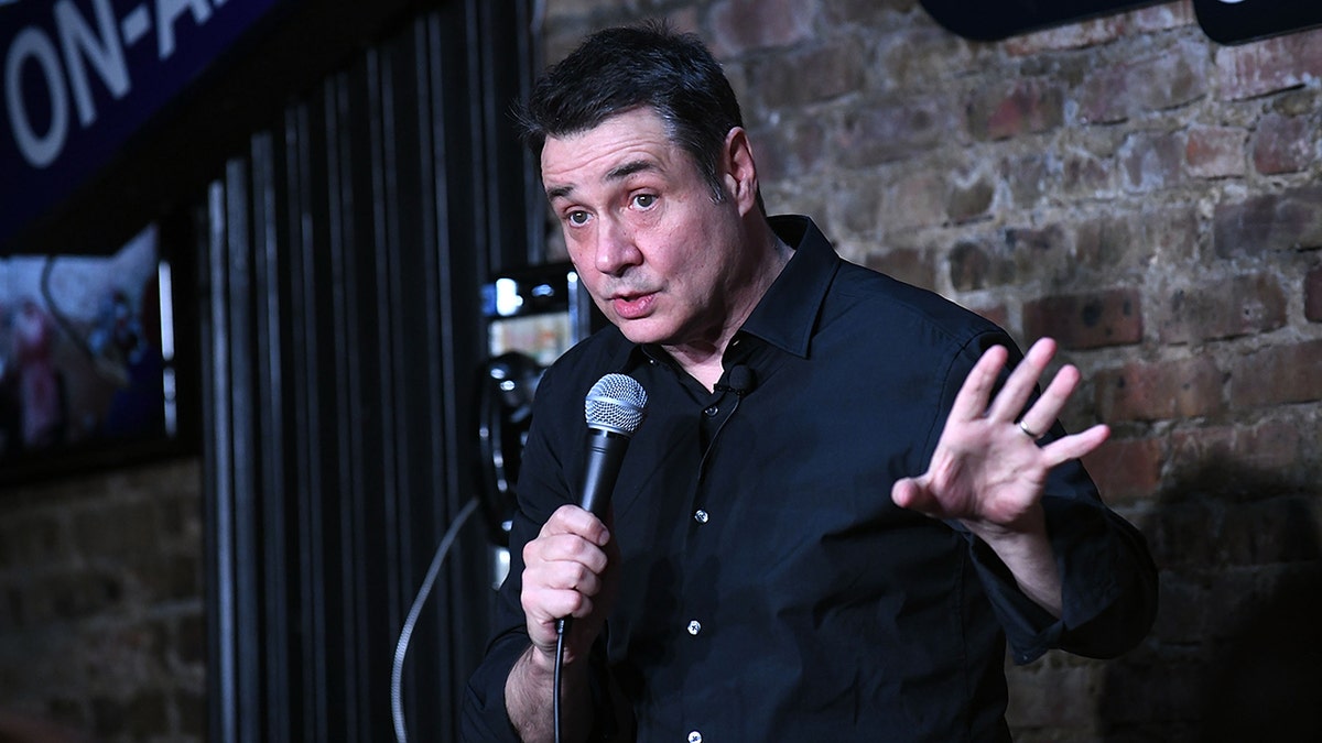 Adam Ferrara in a navy shirt performs on stage holding a microphone