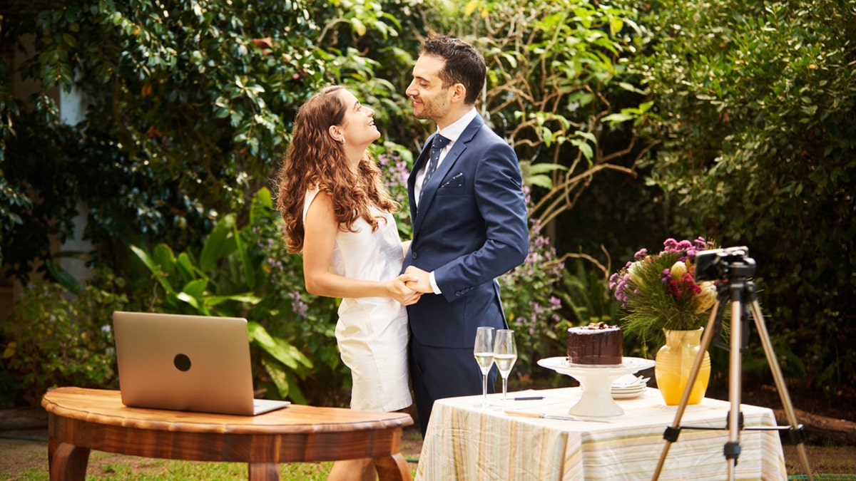 Smiling newlyweds hold hands together while live streaming their outdoor wedding.