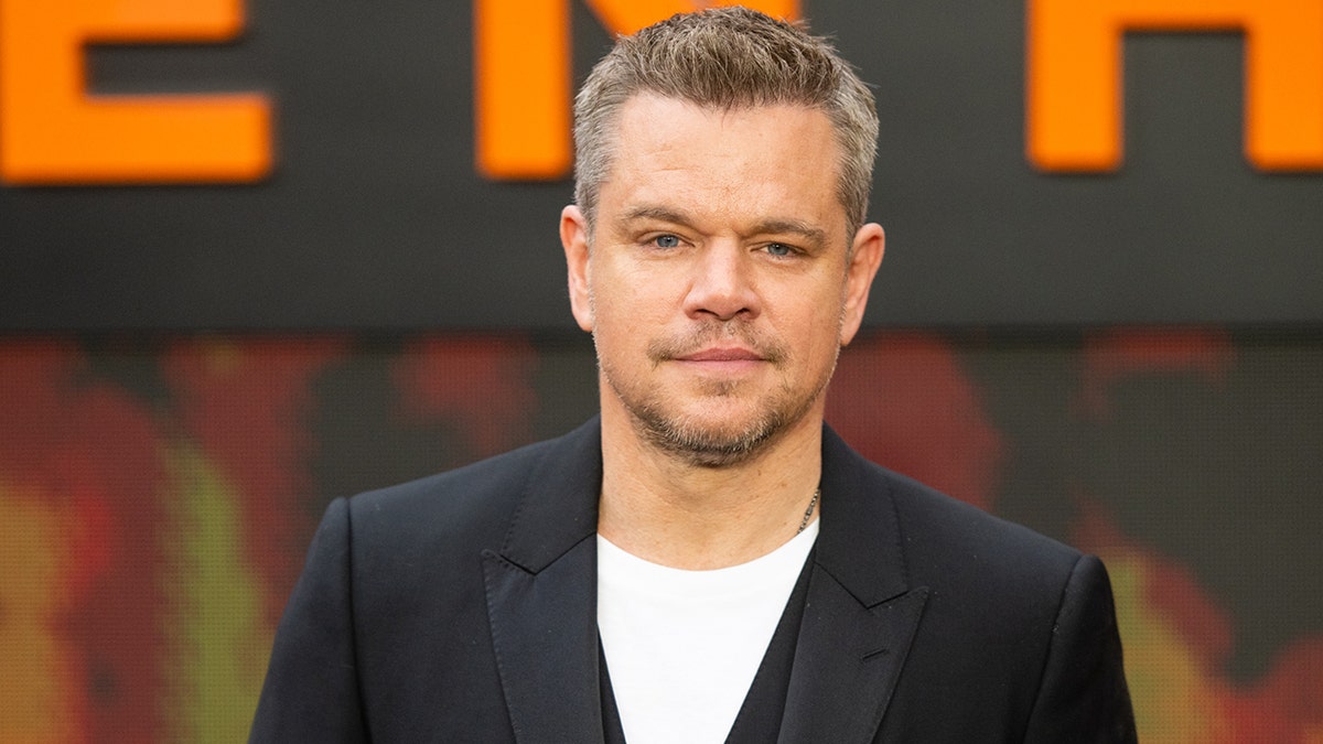 Matt Damon in a black suit gazes directly at the camera on the red carpet