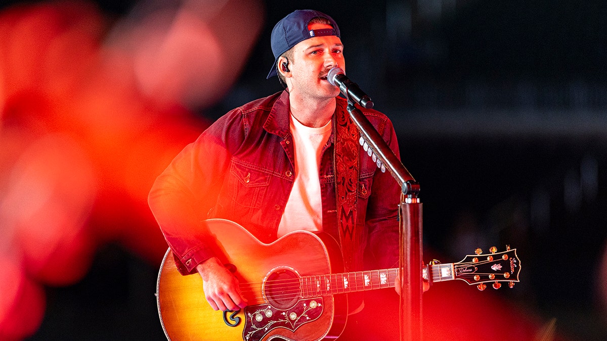 Morgan Wallen performs on stage with a guitar, wearing a red shirt and a backwards hat