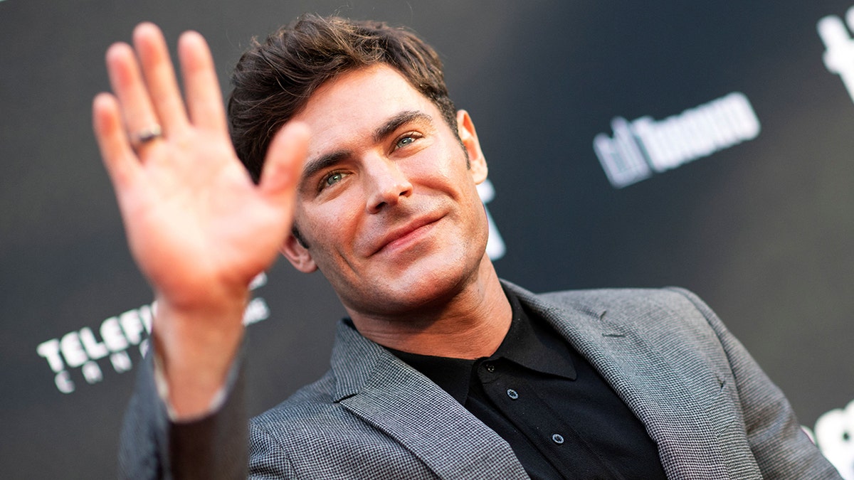 Zac Efron in a black shirt and grey suit looks up as he waves to the crowd at a premiere